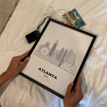 Person holding a framed print of Atlanta with a cityscape design on a bed.