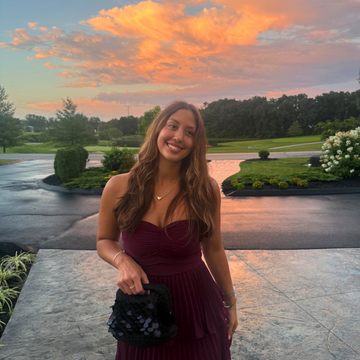 A woman stands outdoors on a stone walkway at sunset wearing a sleeveless burgundy pleated dress and holding a black embellished clutch. Trees, lawn, and a driveway are visible in the background under a dramatic orange sky.