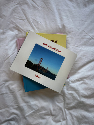 Stack of books with a cover featuring the Golden Gate Bridge and 'San Francisco 2025' text on a white fabric background.